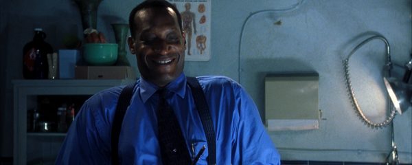 A smiling man wearing a blue shirt with suspenders, showcasing a friendly demeanor in a dimly lit environment.